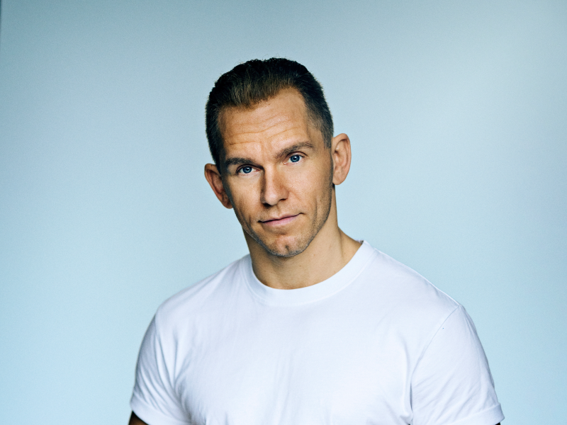 En mand står i en hvid t-shirt på en blå baggrund og kigger ind i kameraet.